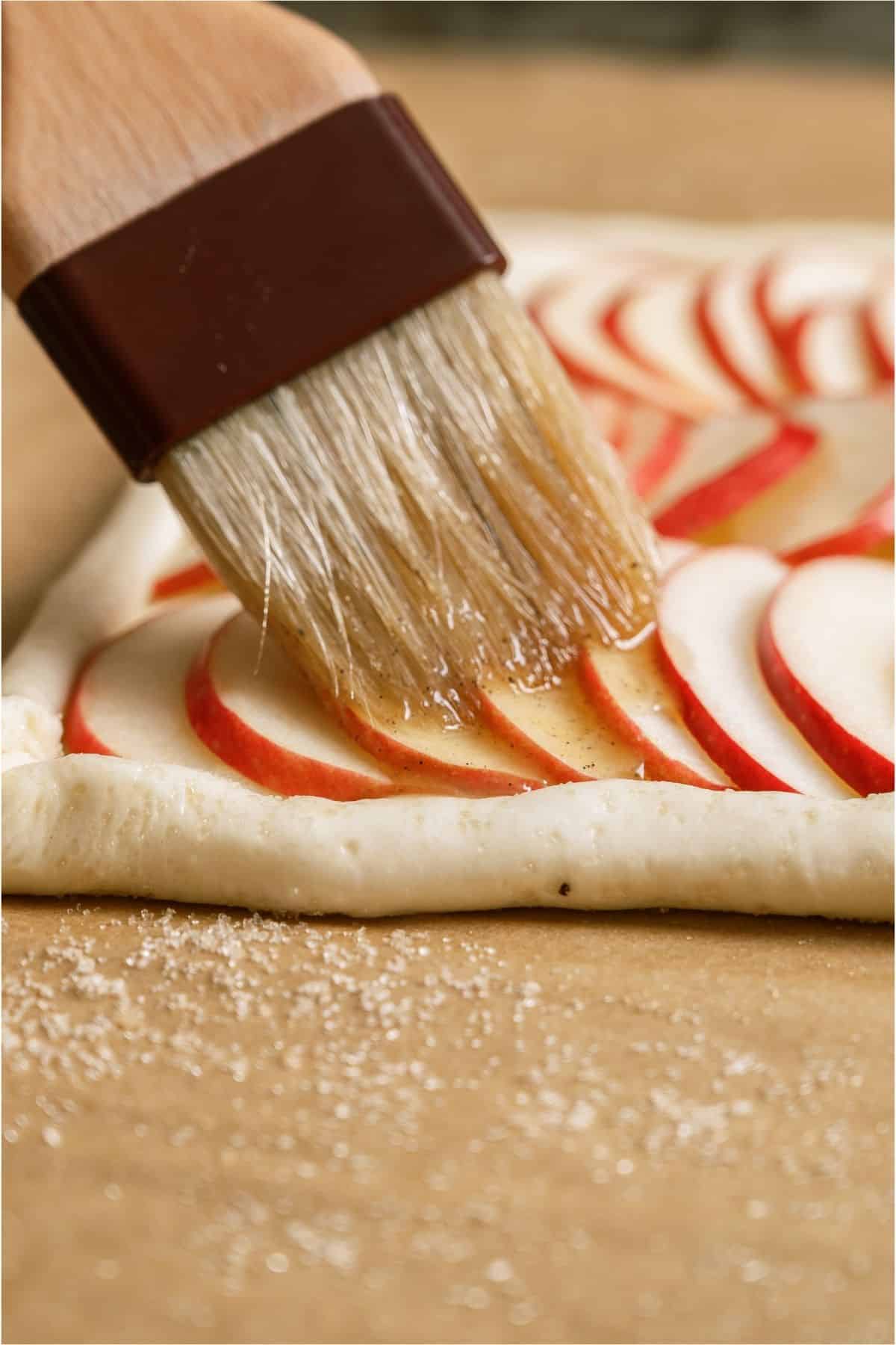 A pastry brush brushing the butter mixture on top. of the sliced apples.