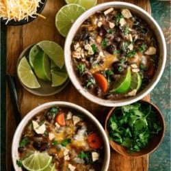 Two bowls of black bean soup with lime wedges, carrots, melted cheese, crushed tortilla chips, and cilantro, served on a wooden board with extra lime, shredded cheese, chips, and cilantro.