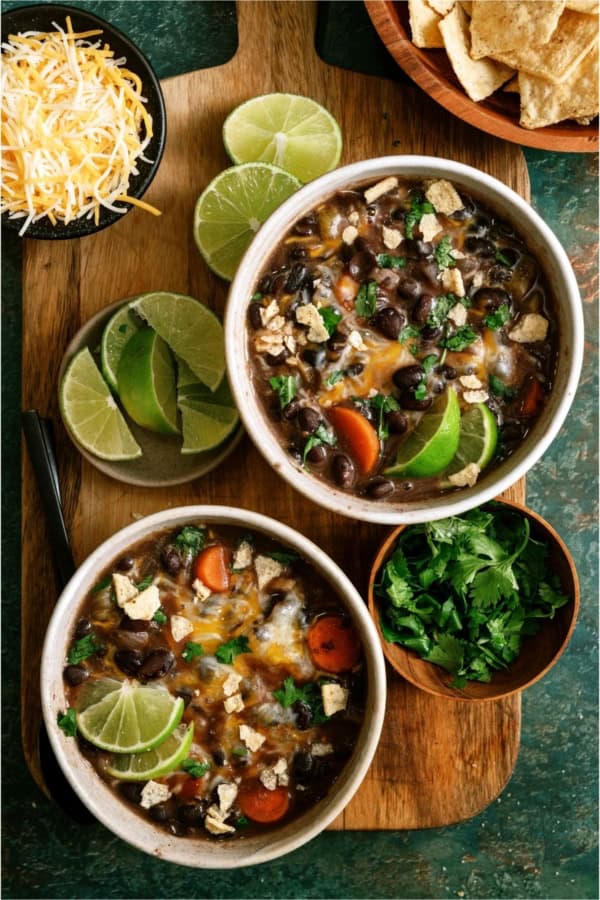 Two bowls of black bean soup with lime wedges, carrots, melted cheese, crushed tortilla chips, and cilantro, served on a wooden board with extra lime, shredded cheese, chips, and cilantro.