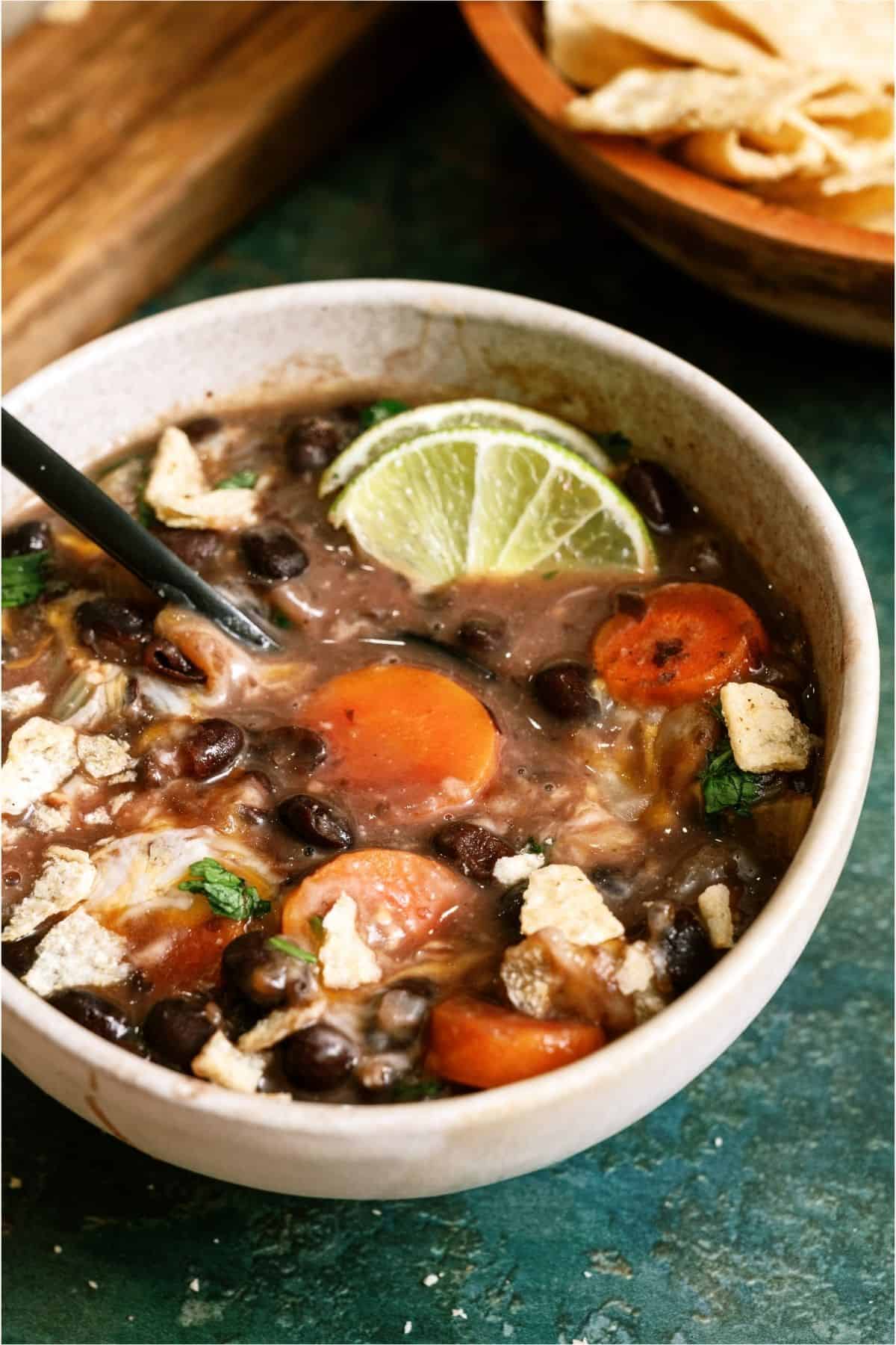 A bowl of black bean soup with carrot slices, tortilla chip pieces, and a lime wedge, with a spoon inside the bowl and tortilla chips in a separate bowl in the background.