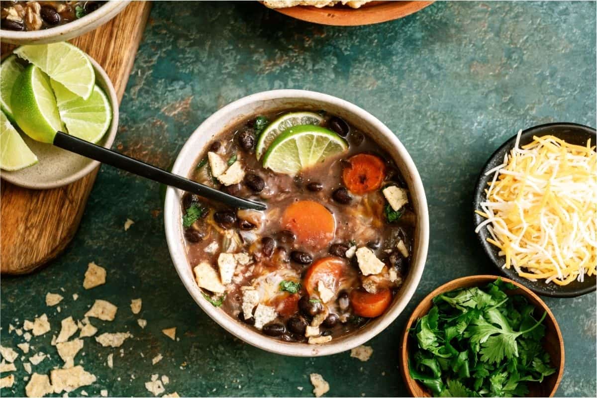 A bowl of black bean soup with carrots, lime wedges, crushed tortilla chips, and a spoon, with shredded cheese and fresh cilantro on the side.