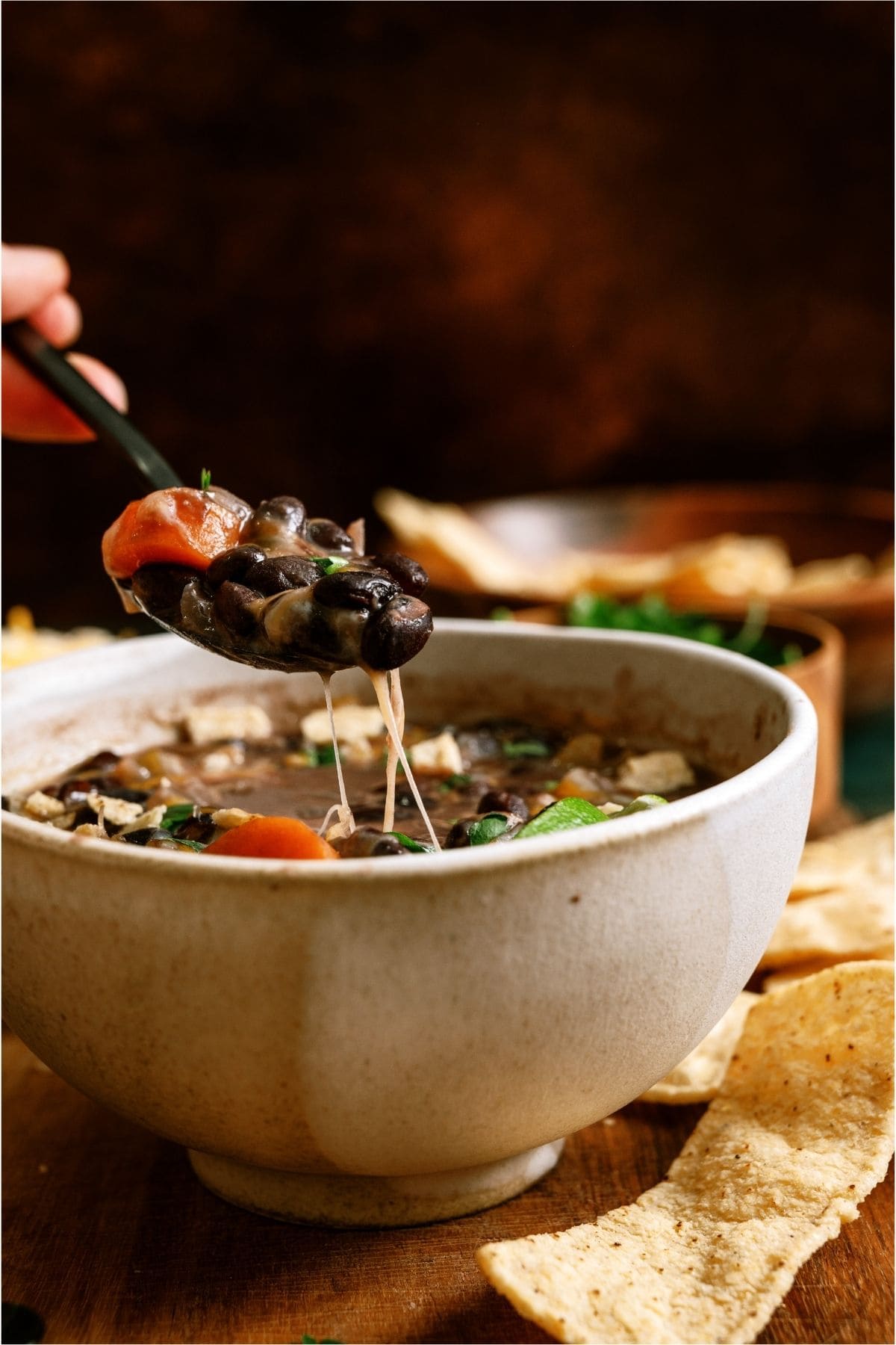A hand holds a spoonful of black bean soup with cheese, vegetables, and herbs above a ceramic bowl; tortilla chips are on the side.