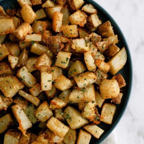 A bowl filled with seasoned, roasted potato cubes, placed on a white surface with part of a cloth visible underneath.