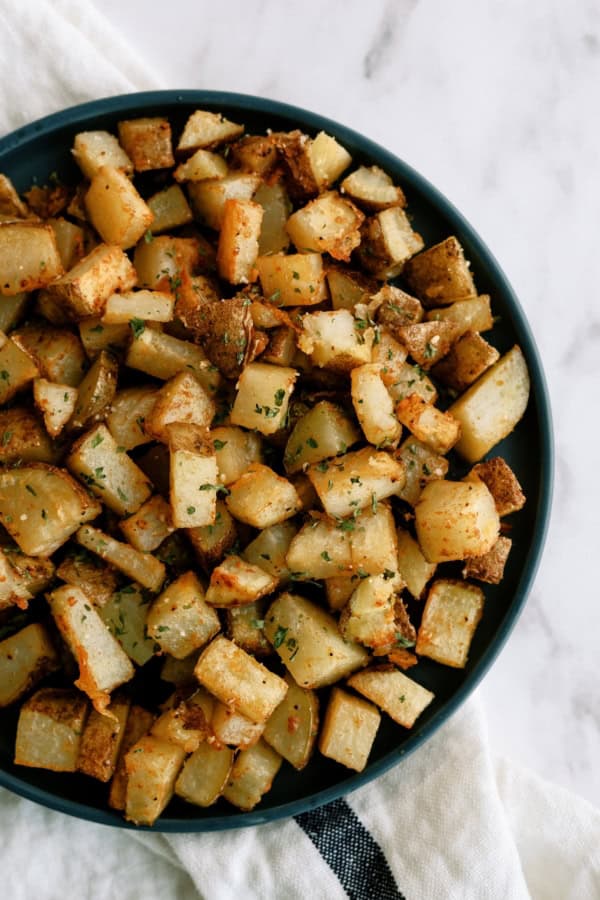 A bowl filled with seasoned, roasted potato cubes, placed on a white surface with part of a cloth visible underneath.
