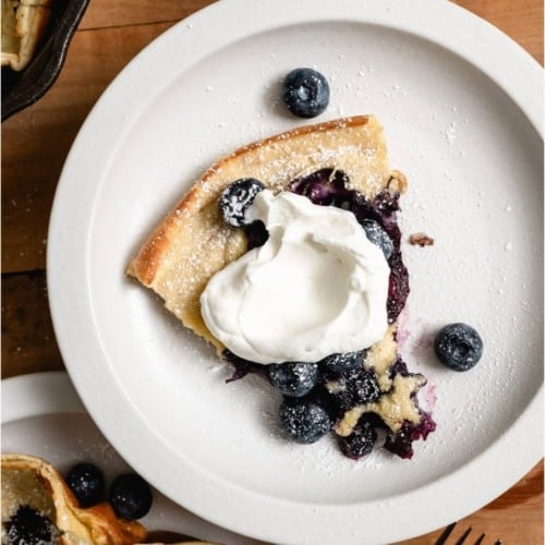 Slice of Dutch Baby on a white plate topped with whip cream and fresh blueberries.
