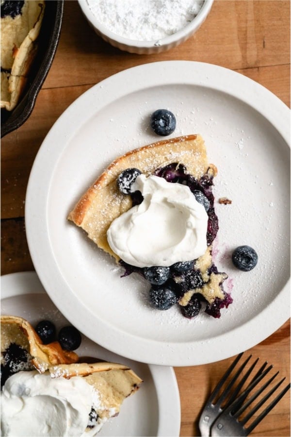 Slice of Dutch Baby on a white plate topped with whip cream and fresh blueberries.