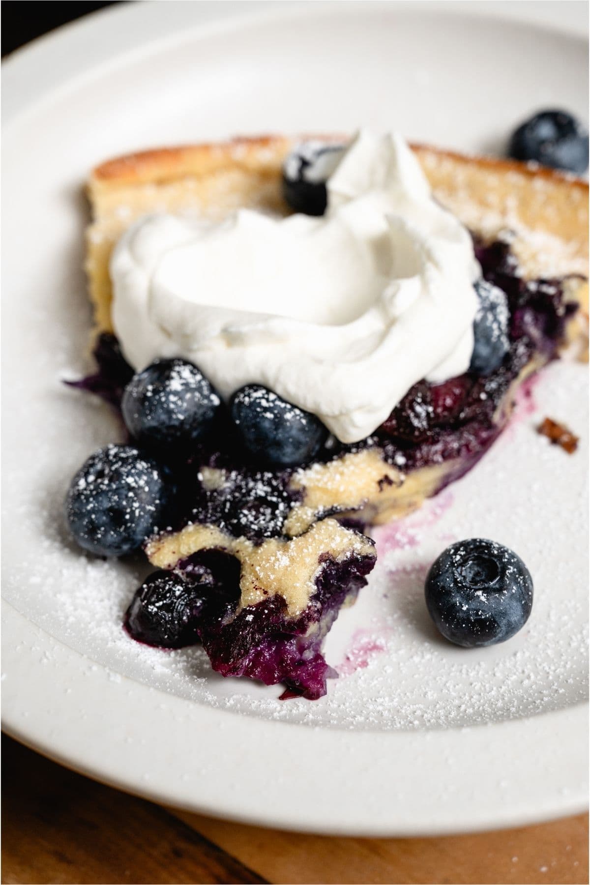 Slice of Dutch Baby on a white plate topped with whip cream and fresh blueberries.