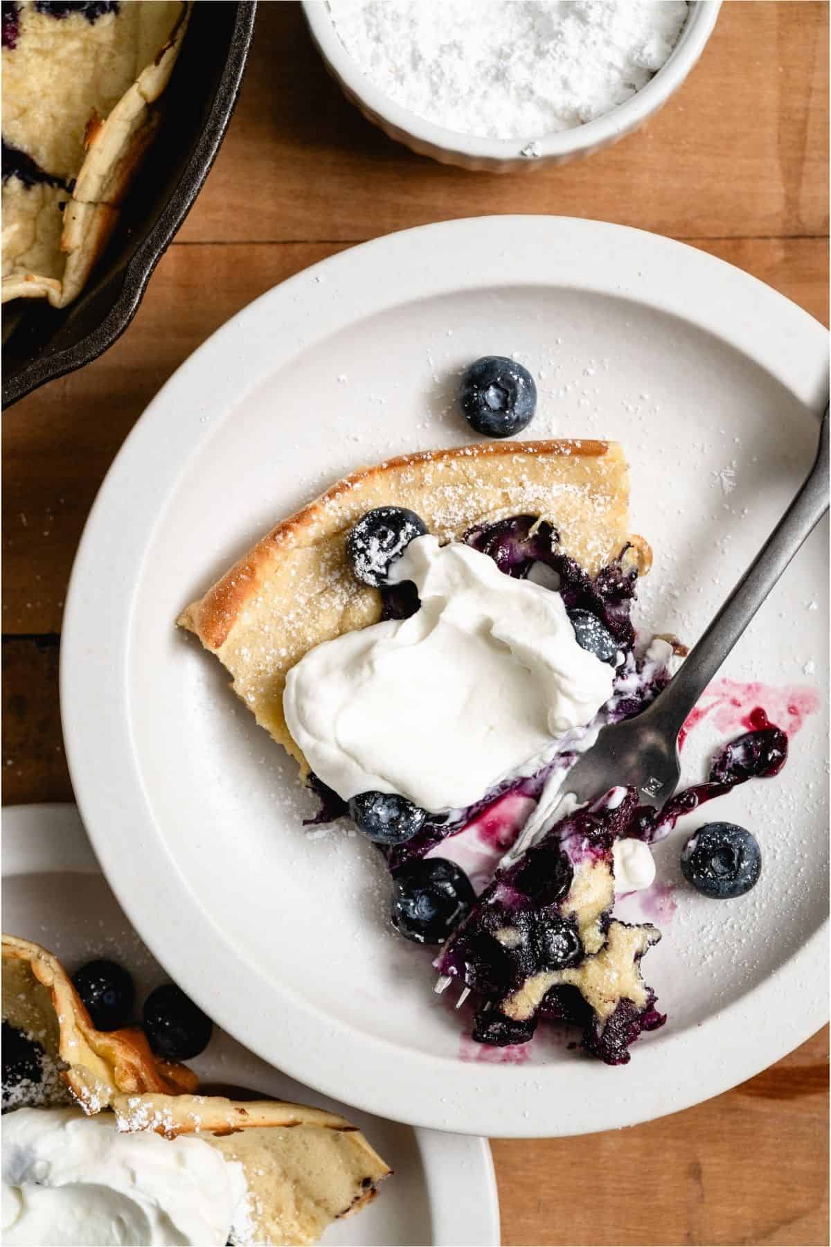 A slice of Dutch Baby on a white plate topped with whip cream and blueberries.