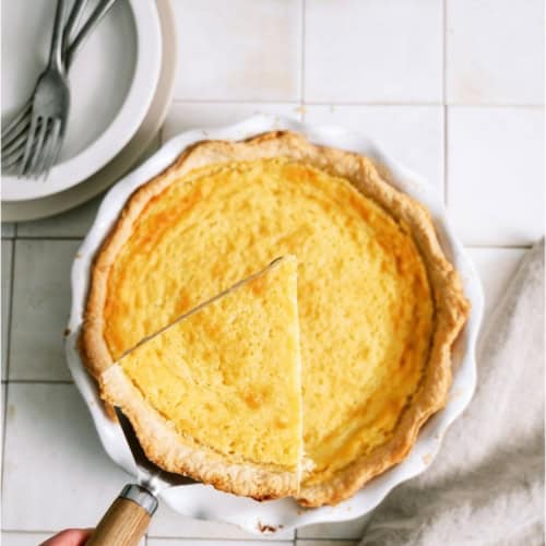 Homemade Buttermilk Pie in a white pie plate with a hand lifting out a slice.