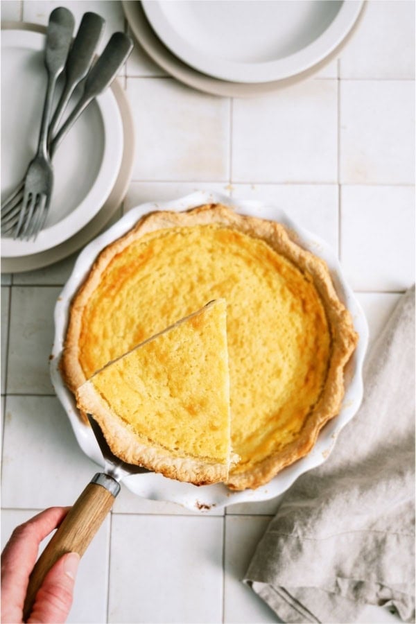 Homemade Buttermilk Pie in a white pie plate with a hand lifting out a slice.