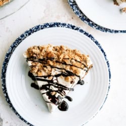 A slice of pie with a crumb topping and chocolate drizzle on a white plate with a blue patterned rim, next to another plate and a partial view of a pie dish.