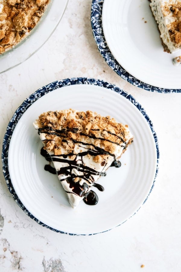 A slice of pie with a crumb topping and chocolate drizzle on a white plate with a blue patterned rim, next to another plate and a partial view of a pie dish.