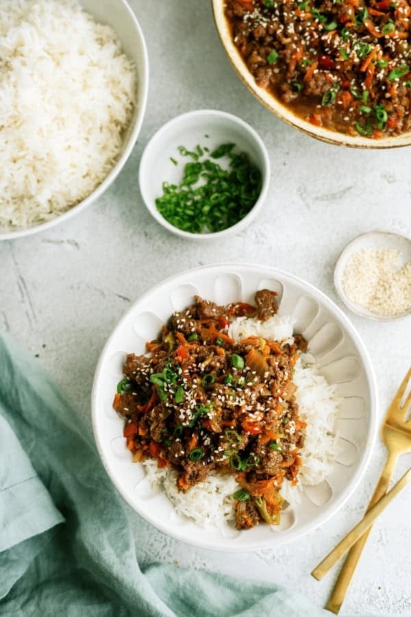 A bowl of white rice topped with stir-fried vegetables and ground meat, garnished with chopped green onions and sesame seeds; bowls of rice, green onions, and sesame seeds nearby.