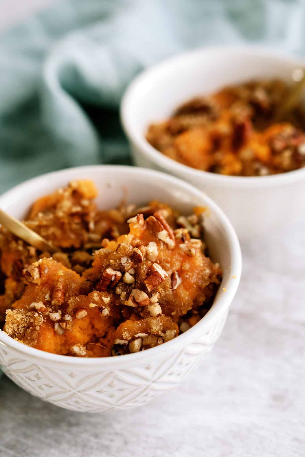 Two white bowls filled with sweet potato casserole topped with chopped pecans and brown sugar, placed on a light surface.