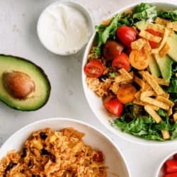 Two bowls with Mexican rice, beans, lettuce, tomatoes, avocado, and tortilla strips, surrounded by small bowls of lettuce, sour cream, and toppings, plus a halved avocado.