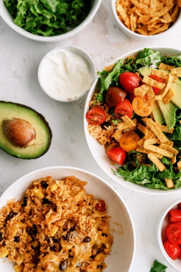 Two bowls with Mexican rice, beans, lettuce, tomatoes, avocado, and tortilla strips, surrounded by small bowls of lettuce, sour cream, and toppings, plus a halved avocado.
