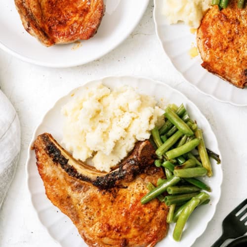 A white plate with a cooked pork chop, mashed potatoes, and green beans. Two additional plates with similar servings are partially visible.