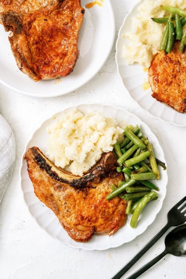 A white plate with a cooked pork chop, mashed potatoes, and green beans. Two additional plates with similar servings are partially visible.