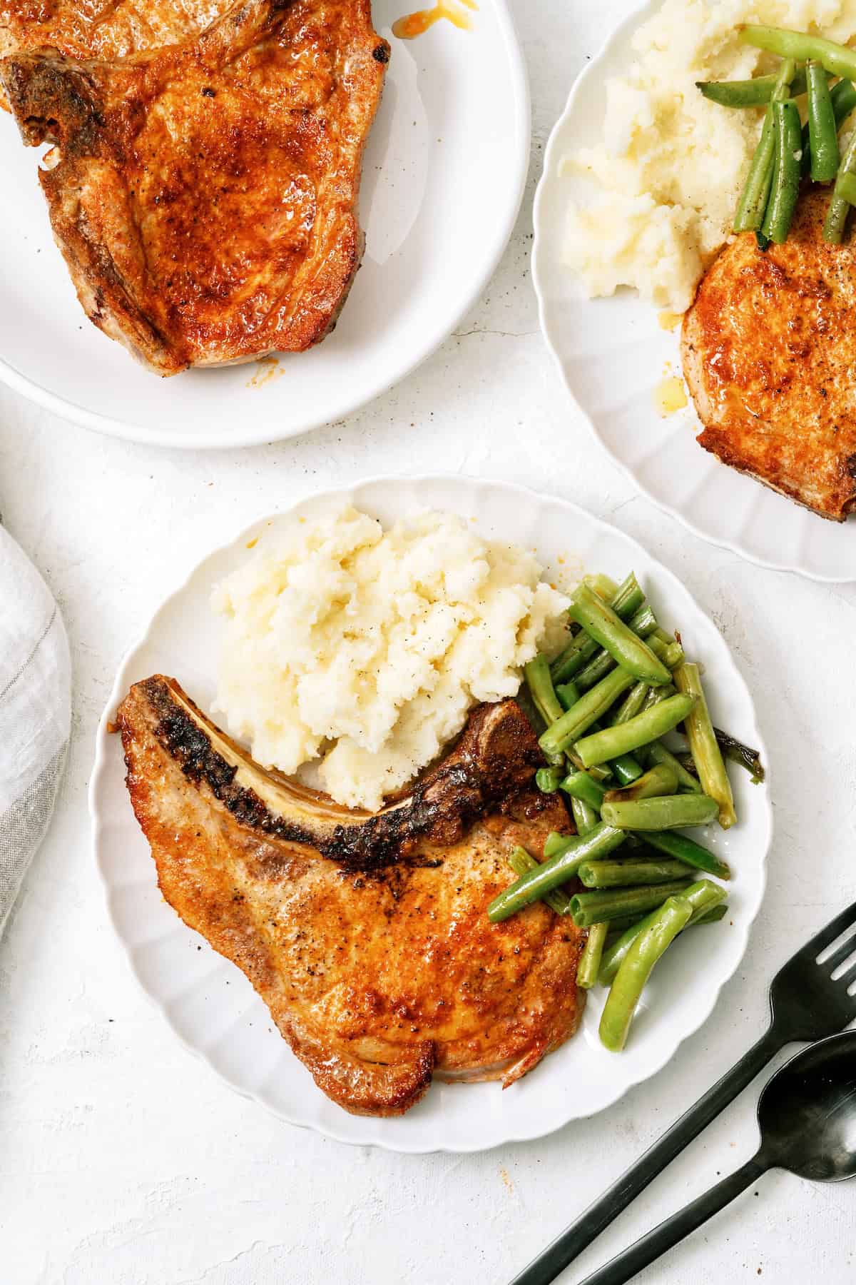 A white plate with a cooked pork chop, mashed potatoes, and green beans. Two additional plates with similar servings are partially visible.