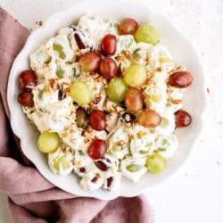 A white bowl of grape salad with red and green grapes, creamy dressing, and chopped nuts, placed on a pink cloth on a white surface.