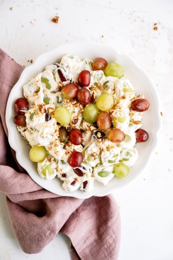 A white bowl of grape salad with red and green grapes, creamy dressing, and chopped nuts, placed on a pink cloth on a white surface.