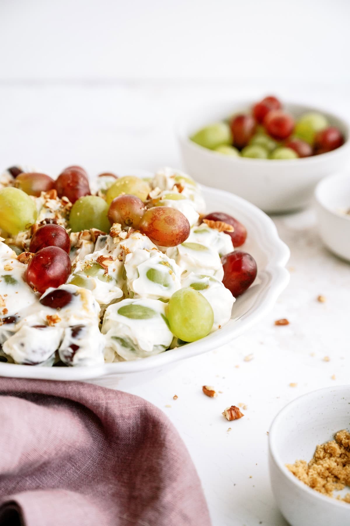 A white plate filled with creamy grape salad topped with chopped nuts, with bowls of grapes and crushed nuts in the background.