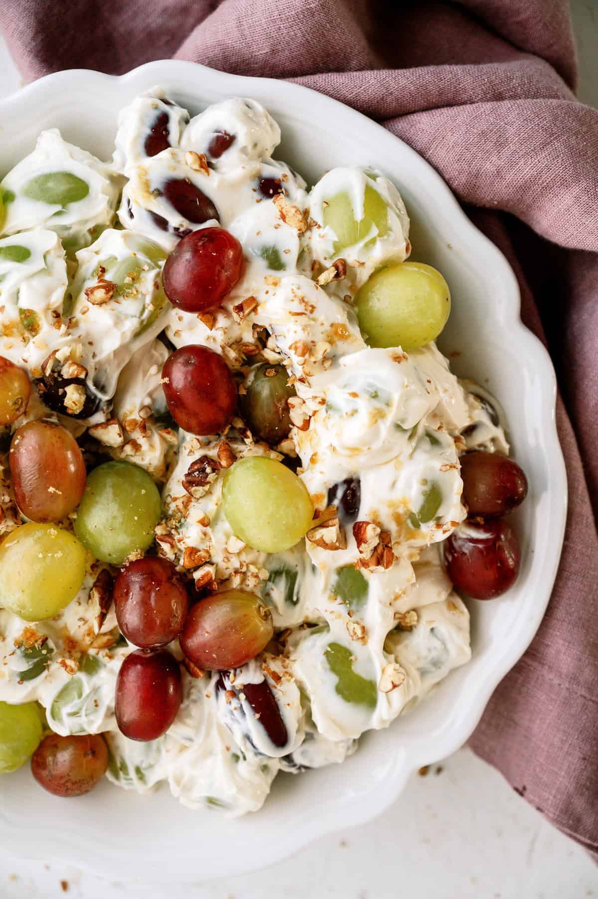 A bowl of grape salad with red and green grapes, creamy dressing, and sprinkled chopped pecans, placed on a mauve napkin.