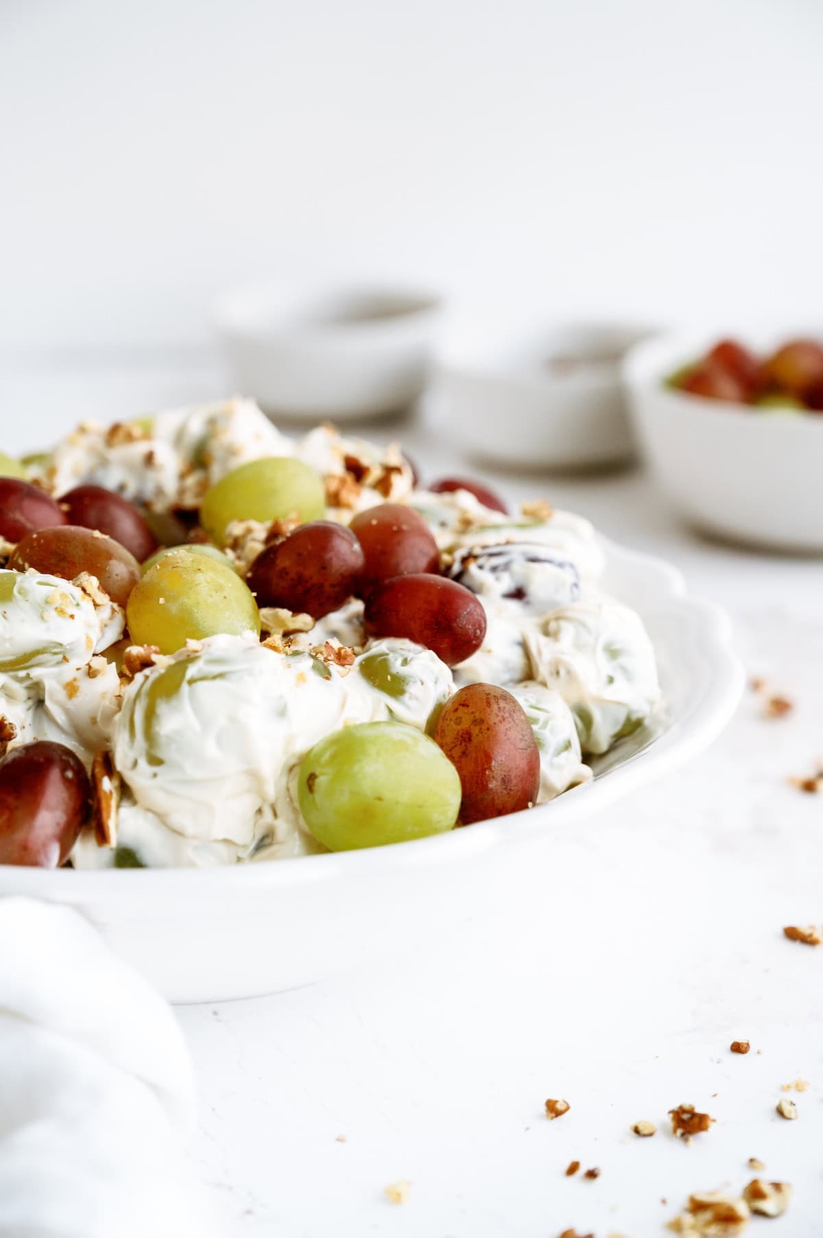 A white bowl filled with green and red grapes coated in a creamy mixture, topped with chopped nuts. Other bowls are blurred in the background.