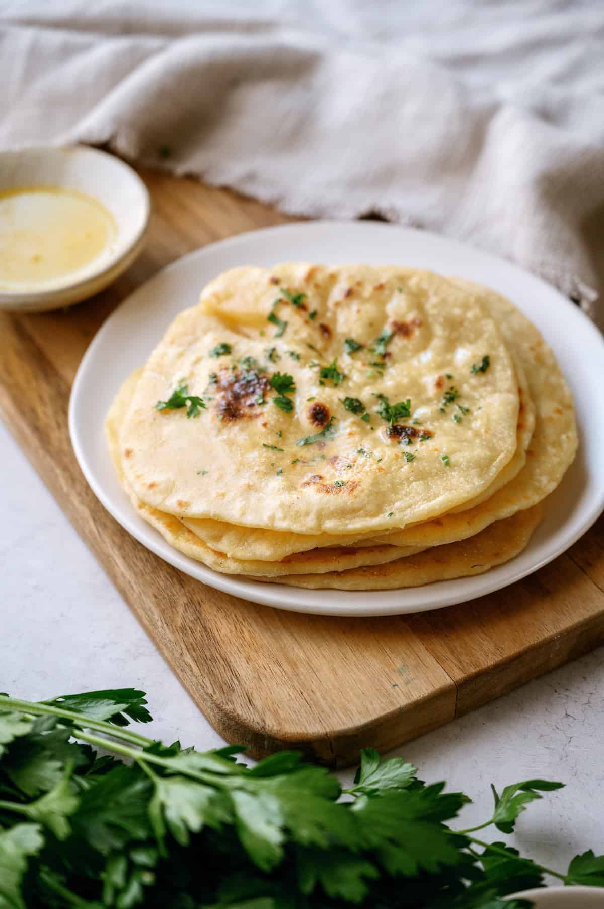 A plate of flatbreads garnished with fresh herbs sits on a wooden board next to a small bowl of sauce and a bunch of parsley.