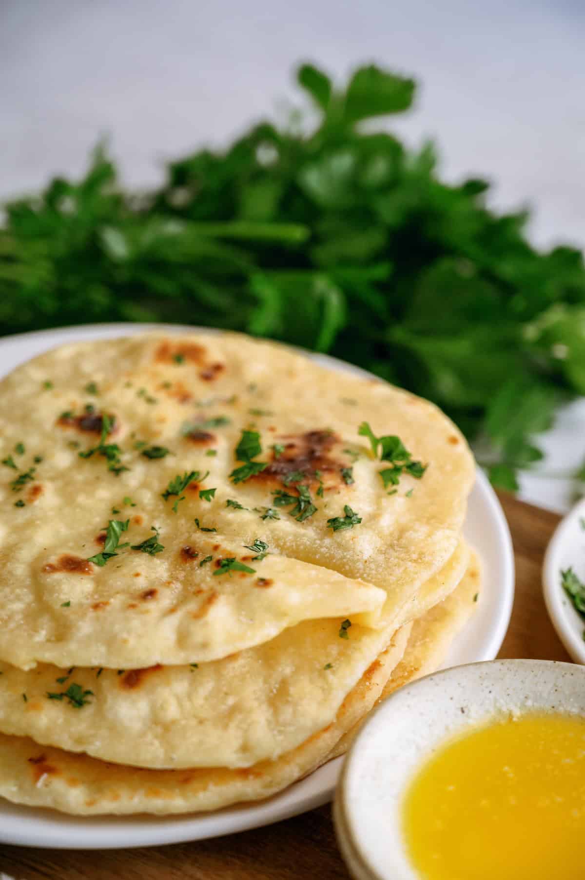 A stack of flatbreads garnished with chopped herbs is on a white plate, with a bowl of melted butter and fresh greens in the background.