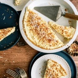 A partially sliced coconut cream pie with toasted coconut topping is served on plates with forks beside it on a wooden table.