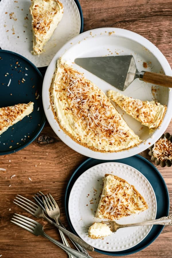 A partially sliced coconut cream pie with toasted coconut topping is served on plates with forks beside it on a wooden table.