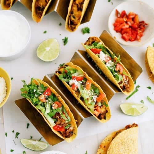 Three hard shell tacos filled with meat, lettuce, tomato, cheese, and cilantro, surrounded by lime wedges, diced tomatoes, chopped cilantro, sour cream, and extra taco shells.