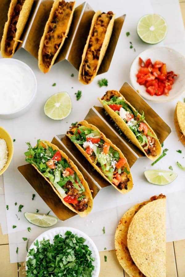 Three hard shell tacos filled with meat, lettuce, tomato, cheese, and cilantro, surrounded by lime wedges, diced tomatoes, chopped cilantro, sour cream, and extra taco shells.