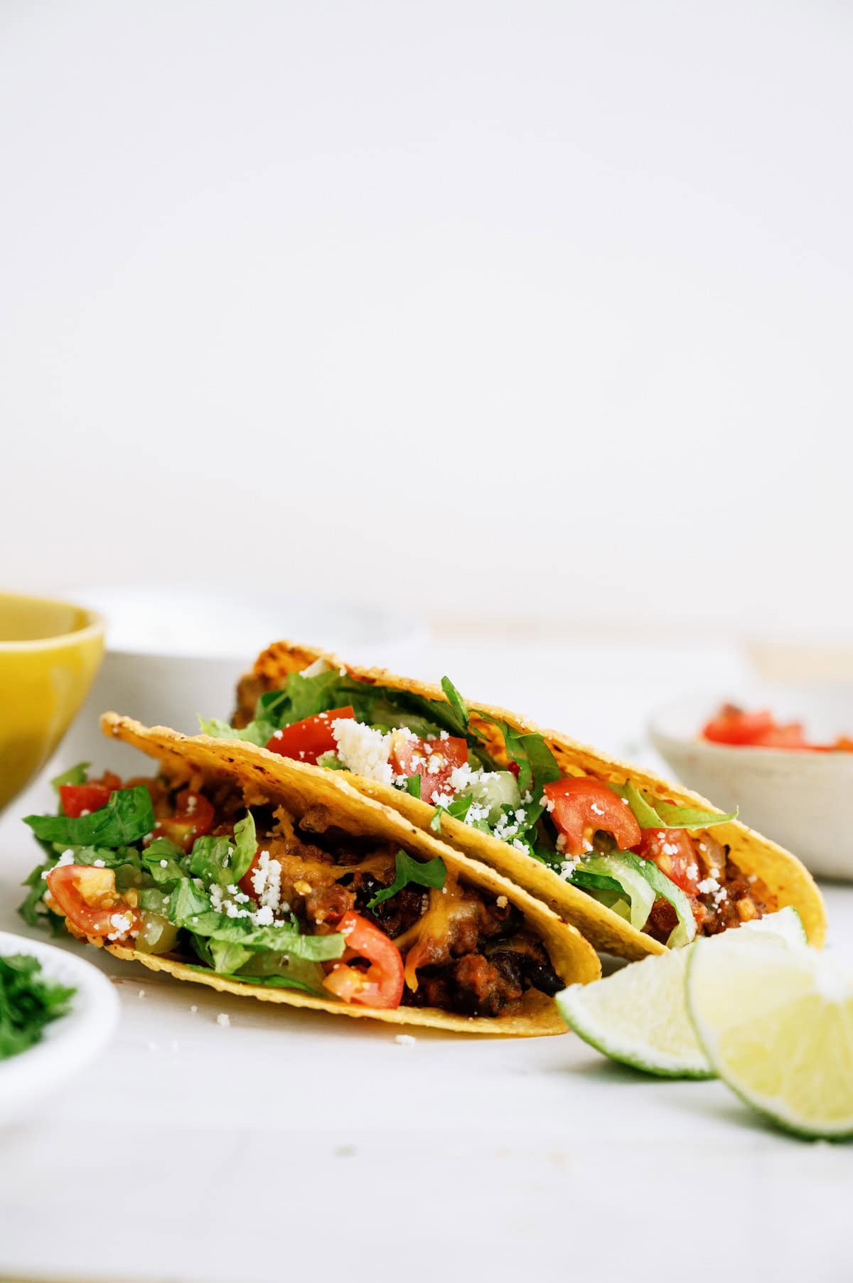 Two hard shell tacos filled with ground beef, lettuce, tomatoes, cheese, and cilantro, with lime wedges on the side.