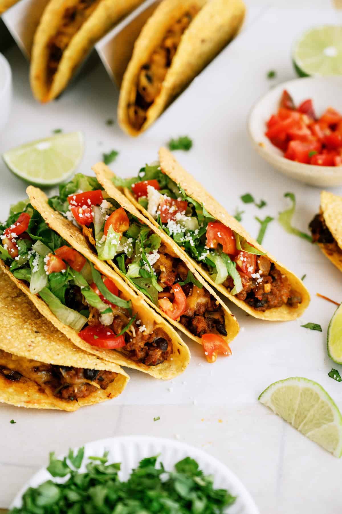 Three hard shell tacos filled with beans, lettuce, tomatoes, and cheese are arranged on a white surface, surrounded by lime wedges, chopped cilantro, and diced tomatoes.