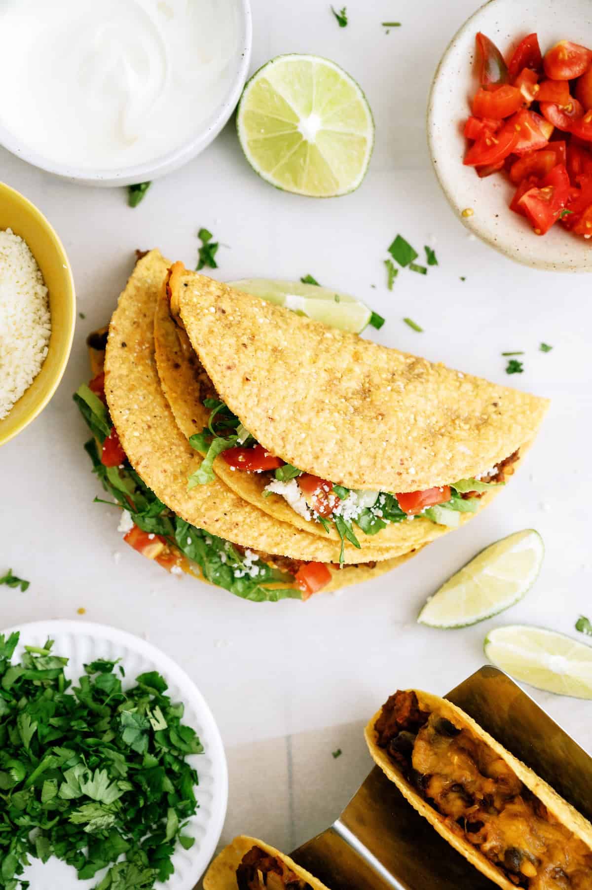 Three hard shell tacos filled with lettuce, tomato, and cheese are on a white surface, surrounded by lime wedges, chopped cilantro, sour cream, and diced tomatoes.