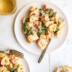 Two plates of creamy tortellini pasta with spinach and tomatoes, served with forks. Two glasses of white wine and a piece of bread are on a white surface nearby.