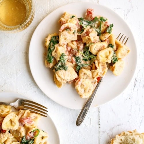 Two plates of creamy tortellini pasta with spinach and tomatoes, served with forks. Two glasses of white wine and a piece of bread are on a white surface nearby.
