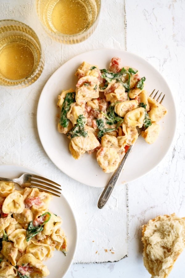Two plates of creamy tortellini pasta with spinach and tomatoes, served with forks. Two glasses of white wine and a piece of bread are on a white surface nearby.