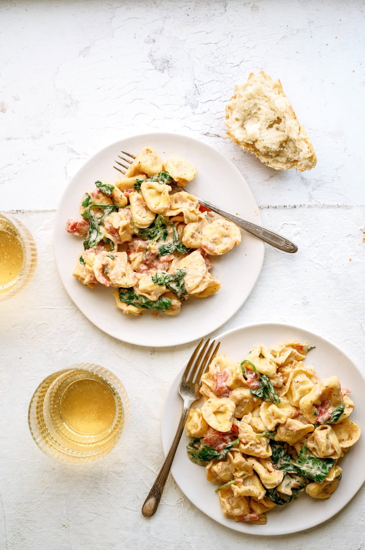 Two plates of creamy tortellini pasta with spinach and tomatoes, each with a fork, are on a white surface next to two glasses of white wine and a piece of bread.