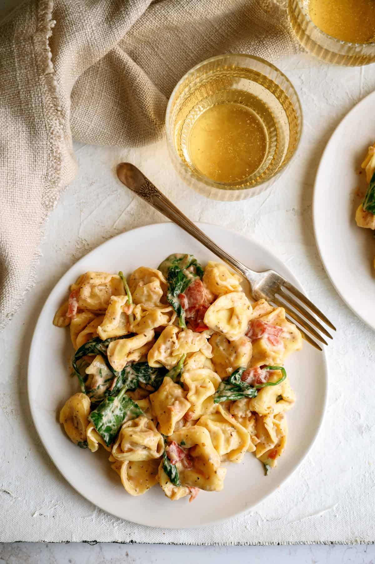 A plate of creamy tortellini pasta with spinach and tomatoes, served with a fork, next to a glass of white wine on a light-colored table.