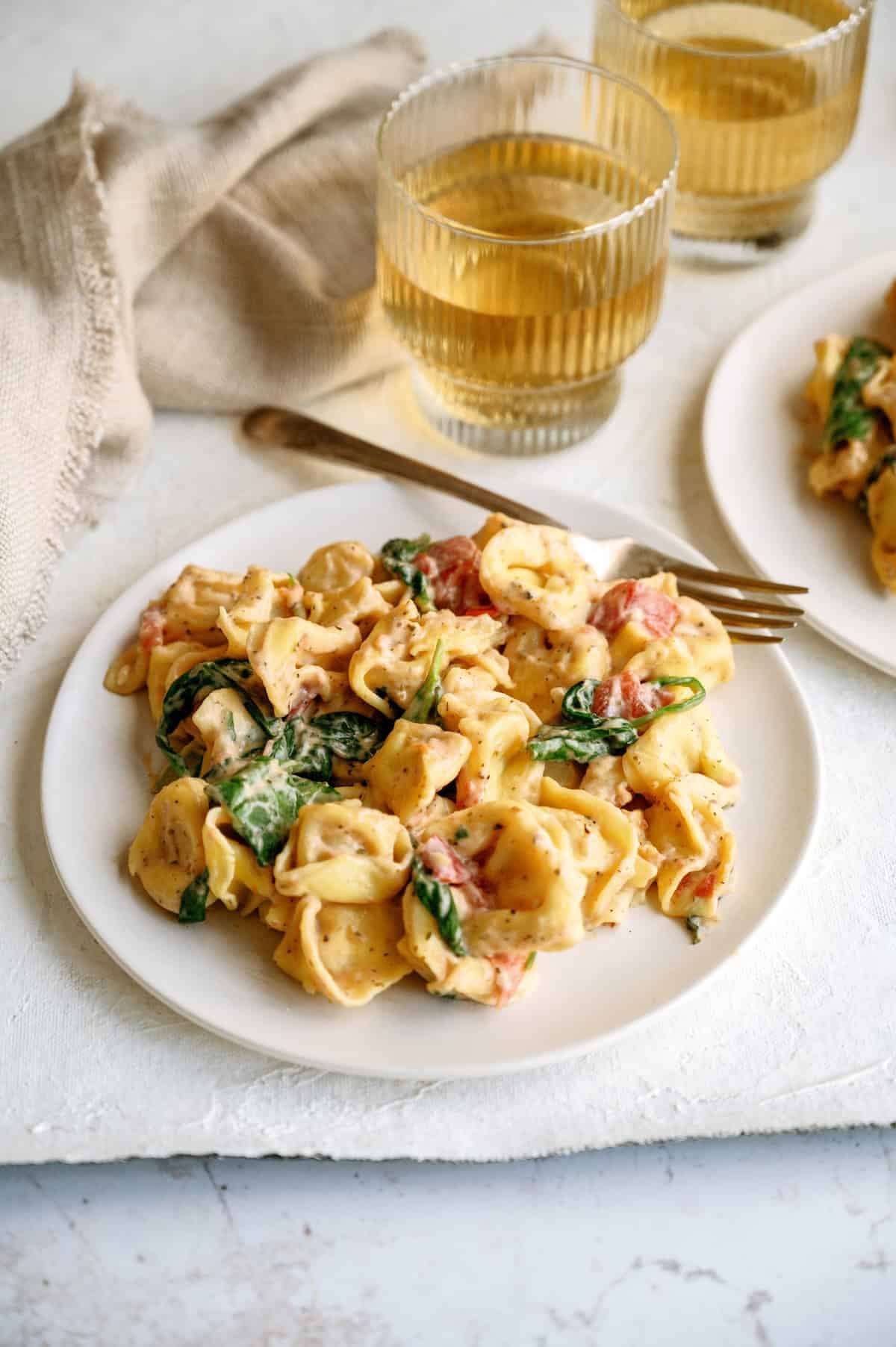 A plate of creamy tortellini pasta with spinach and tomatoes, served with a gold fork and two glasses of light-colored beverage in the background.