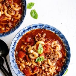 A blue bowl filled with macaroni, ground beef, tomato chunks, and broth, garnished with fresh basil. A croissant and black utensils are beside the bowl.