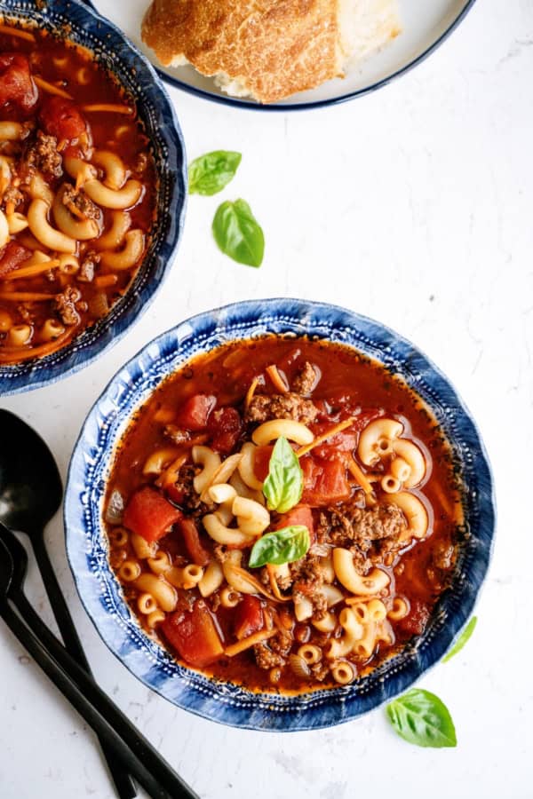 A blue bowl filled with macaroni, ground beef, tomato chunks, and broth, garnished with fresh basil. A croissant and black utensils are beside the bowl.