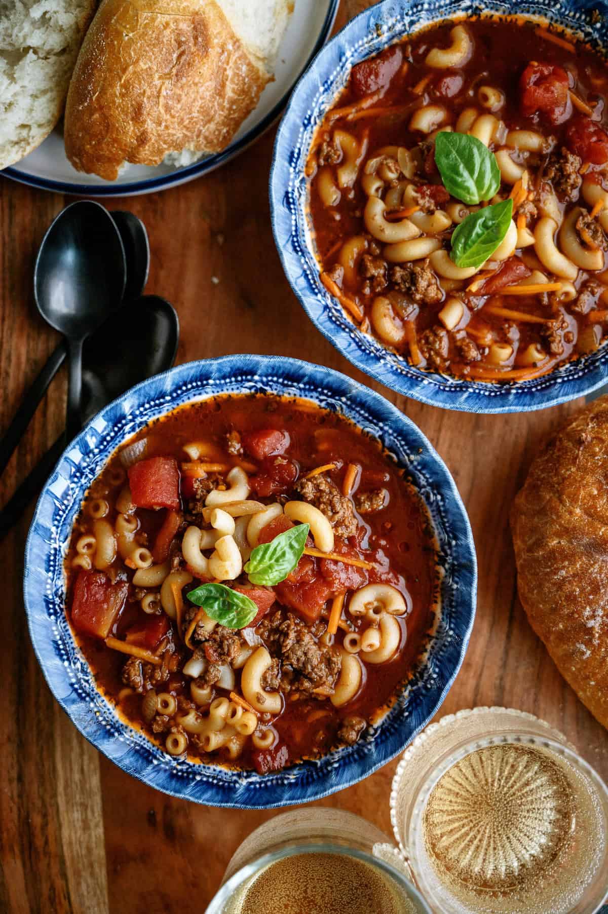 Two bowls of macaroni and beef soup garnished with basil, served with bread on a wooden table beside spoons and a glass of drink.