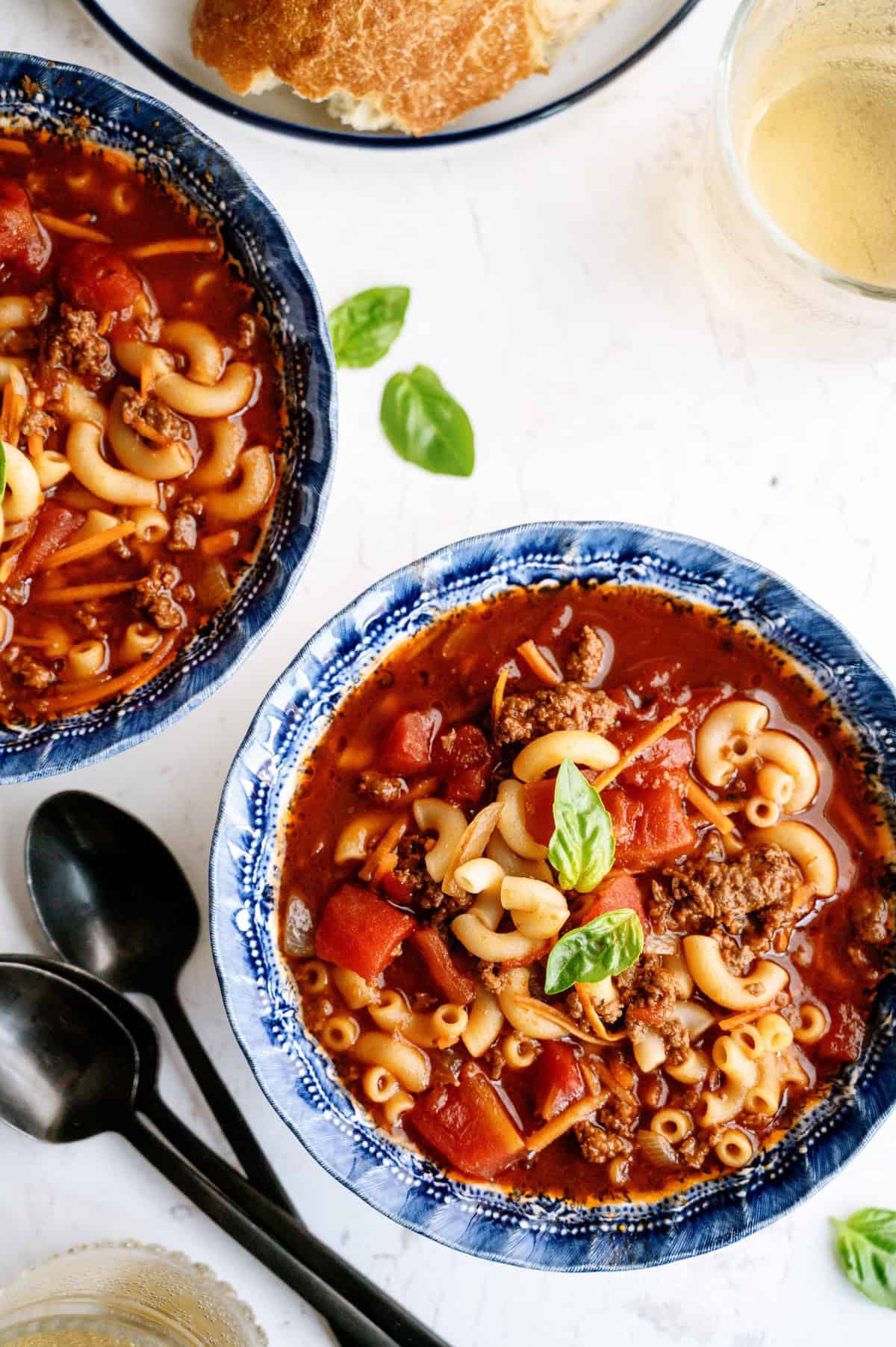 Two bowls of macaroni and beef soup with tomatoes, garnished with basil, next to spoons and a plate with bread on a white surface.