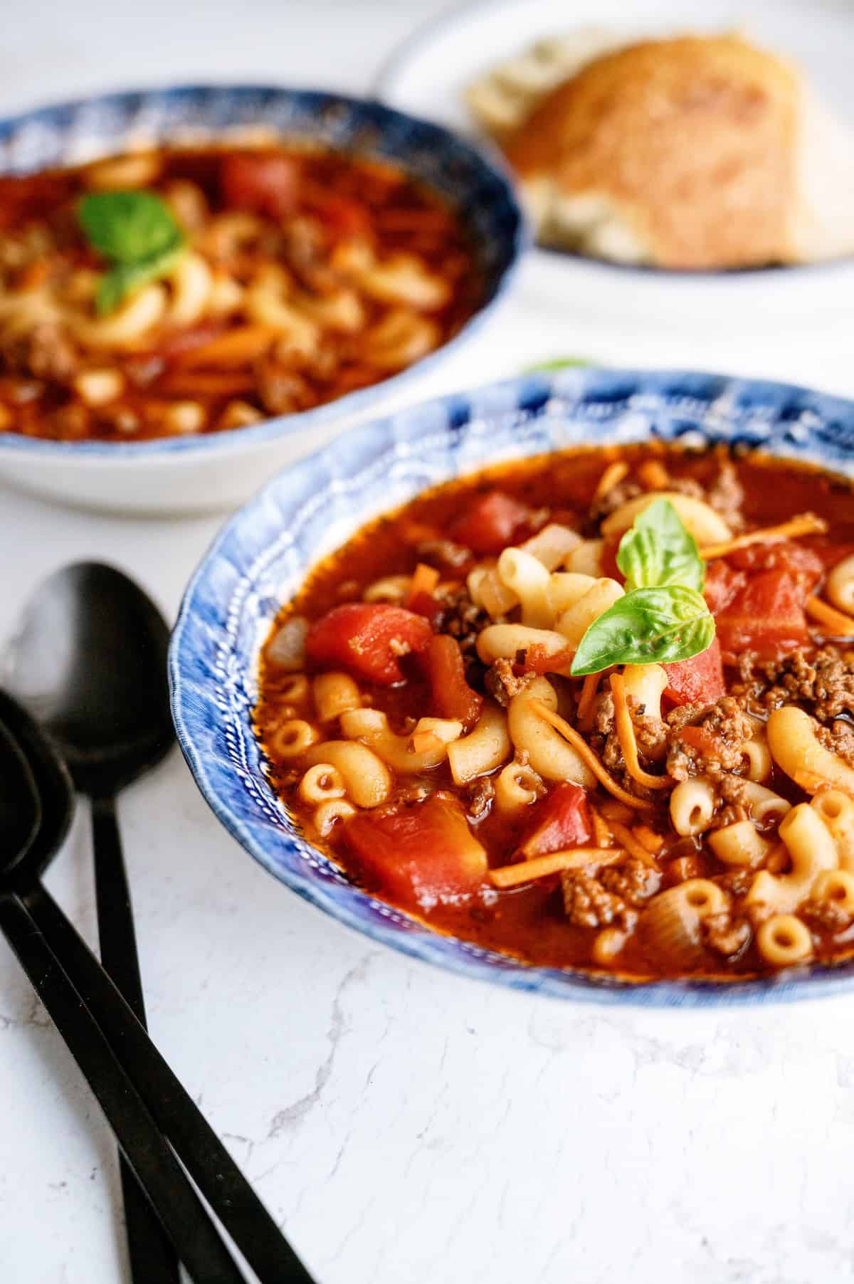 Two bowls of pasta soup with ground meat, tomatoes, and macaroni noodles, topped with basil, are on a white surface with a bread roll and black cutlery beside them.