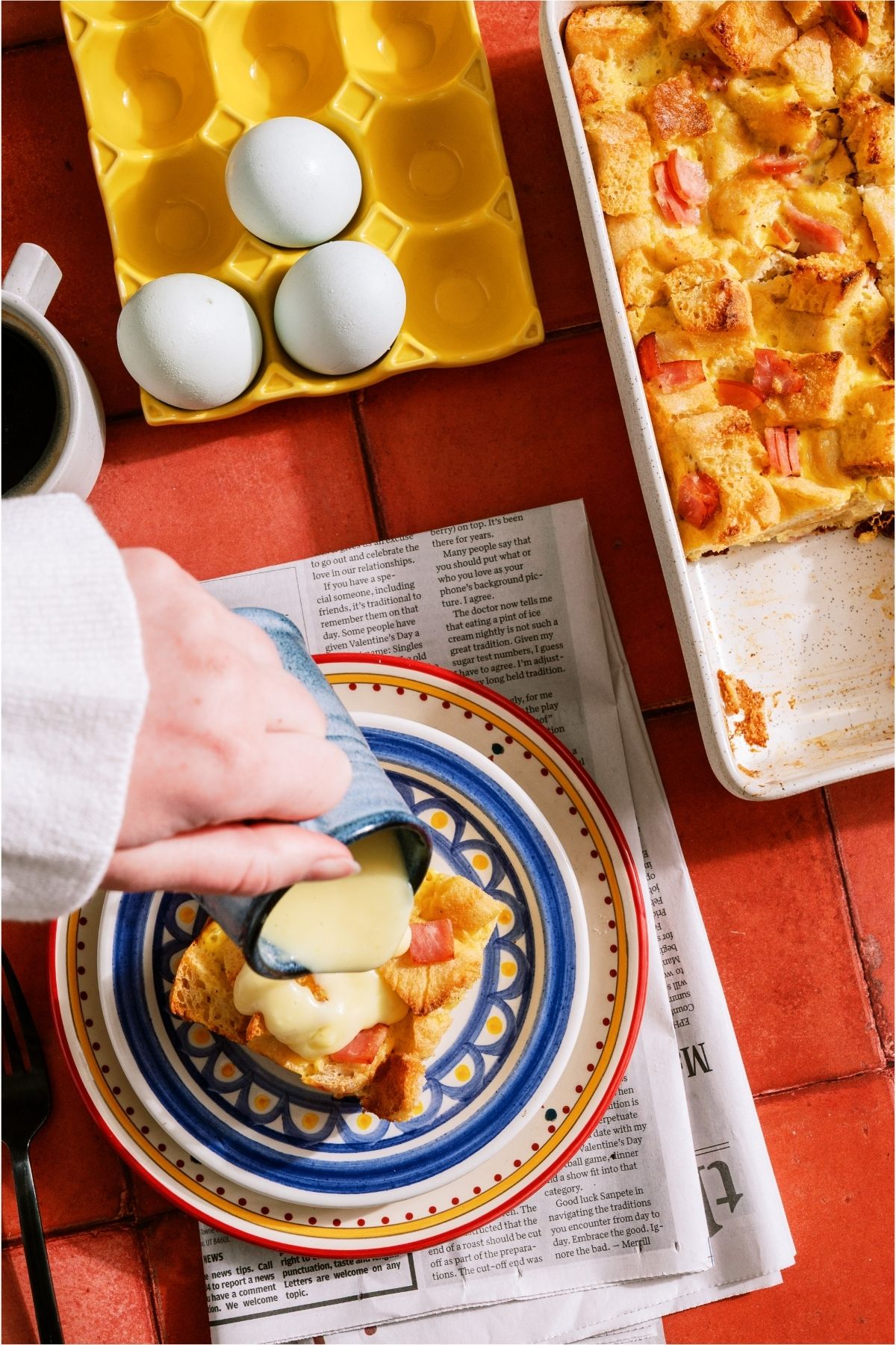 Pouring Hollandaise sauce on top of a slice of Eggs Benedict Casserole on a plate.