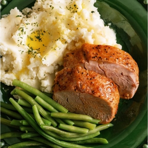 Two slices of Oven Baked Pork Tenderloin on a plate with mashed potatoes and green beans.